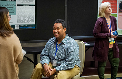 A poster presenter at the AUCD Conference smiles while sitting in front of his poster, talking to another attendee.